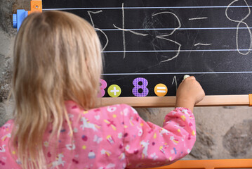 Blond hair girl writing with a chalk on the black board and playing with magnetic numbers.