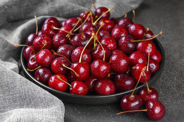 Close up of black plate filled with ripe red sweet cherry placed on gray linen napkin on dark background. Delicious food, juicy summer fruit and healthy eating.