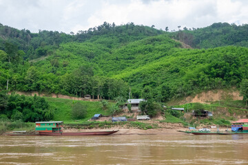 Fototapeta premium Rural village along Mekong River in Laos with boats and green hills.