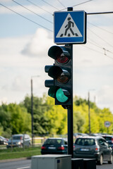 Traffic light showing green signal near pedestrian crossing sign on busy city street with cars in background on a sunny day