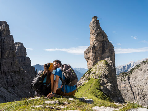 Iconic friulan mountain Campanile Di Val Montanaia, Dolomiti Friulane, Val Cimoliana, Friuli, ItalyA couple with backpacks kisses on a grassy ridge, with the dramatic mountain peak in the background.