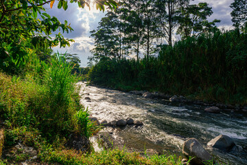 Serene river flowing through lush Colombian landscape with rocks, grasses and cloudy sky.