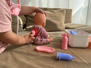 Little girl listens to a doll’s chest using a toy stethoscope. Playing doctor to learn care and...