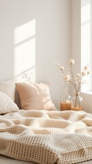 Cozy indoor relaxation scene with soft beige pillows, chunky knit throw blanket, clear vases, and delicate decorative flower branches by a bright window