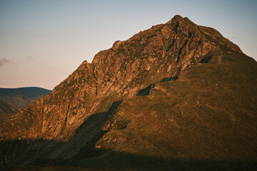 Majestic mountain peak bathed in the golden light of sunset.  Rugged terrain and dramatic shadows create a stunning natural landscape. Ideal for travel, adventure, and nature themes.
