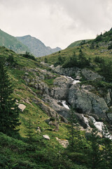 Fototapeta premium Serene mountain landscape with cascading waterfall. Lush green hillsides and rugged grey rocks create a breathtaking natural scene. Perfect for travel, nature, and adventure themes.