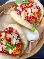 Delicious bao buns filled with colorful vegetables, topped with fresh herbs, chili slices, and coconut flakes, served in a wooden bowl with parchment paper for a vibrant culinary experience