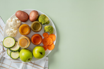 Healthy baby food assortment with fresh vegetables and fruit on a white plate. Includes potatoes, broccoli, cauliflower, zucchini, carrot, green apple, and jars of puree.