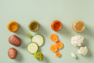 Four jars of baby vegetable puree with matching ingredients arranged in front - potato, zucchini, broccoli, carrot on a light background