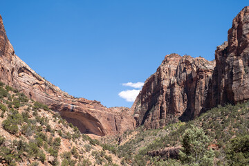 A narrow desert canyon framed by towering sandstone cliffs opens under a bright blue sky, highlighting Zion&rsquo;s dramatic terrain and the immense vertical scale of its rock walls