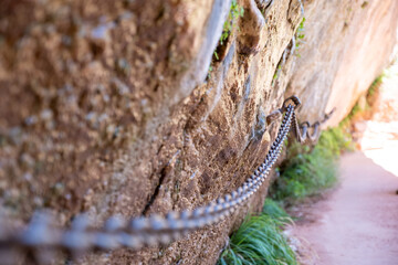 Focus shifts along a taut chain railing leading hikers safely along a narrow rock ledge in Zion, with shallow depth of field emphasizing the steep descent beyond the secured path