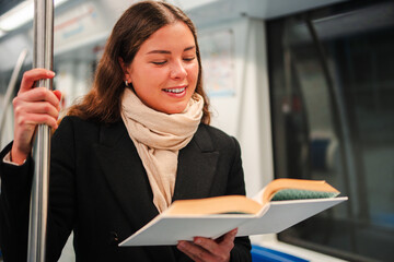 A focused female passenger is deeply engaged in reading a fascinating book on the subway. She is...