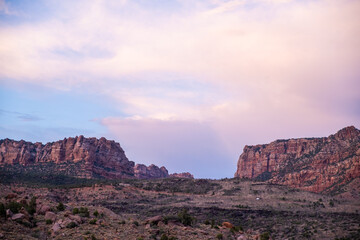 Clouds roll over jagged red rock formations outside Zion National Park as late sunlight illuminates the foreground with pastel hues