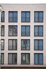 Beige brick building facade with four rows of evenly spaced windows, each window framed with black metal railings. Reflective glass mirrors surrounding architecture under daylight.

