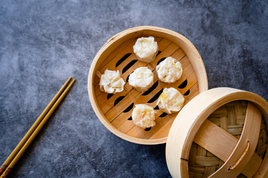 Top-down view of traditional steamed dumplings arranged in a bamboo steamer with a lid and wooden chopsticks on a textured gray surface. A classic presentation of Asian cuisine.