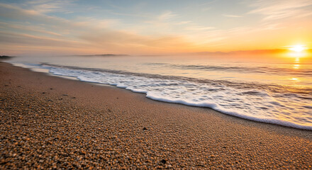 A tranquil beach at sunrise, with soft golden light illuminating a misty horizon and gentle waves lapping the sandy shore