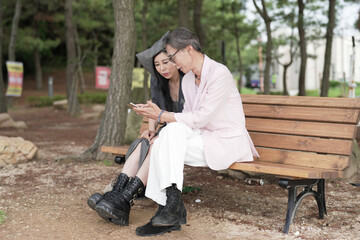 Fototapeta premium In June 2025, a Korean man and woman in their late fifties sit on a bench by the sea, chatting and smiling as they enjoy the breeze, ocean view, and calm surroundings in Incheon Metropolitan City.