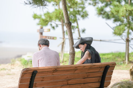 In June 2025, a Korean man and woman in their late fifties sit on a bench by the sea, chatting and smiling as they enjoy the breeze, ocean view, and calm surroundings in Incheon Metropolitan City. - Powered by Adobe