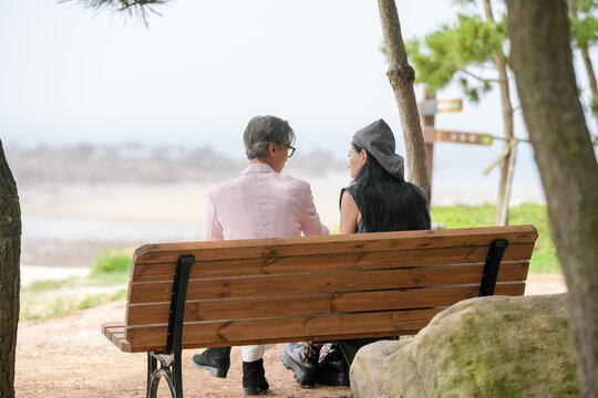In June 2025, a Korean man and woman in their late fifties sit on a bench by the sea, chatting and smiling as they enjoy the breeze, ocean view, and calm surroundings in Incheon Metropolitan City.