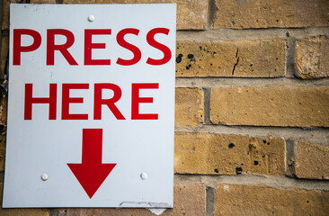 Large "PRESS HERE" sign in bold red letters with a downward-pointing red arrow, mounted on a brick wall. Attention-grabbing signage, possibly for emergency or interactive use.