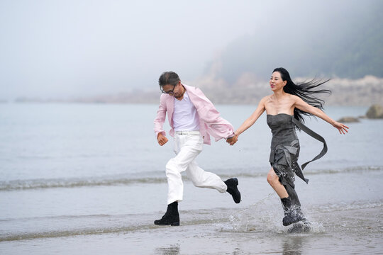 In June 2025, a Korean man and woman in their late fifties are running at full speed along the rugged shoreline filled with jagged rocks and crashing waves in Incheon Metropolitan City, South Korea.