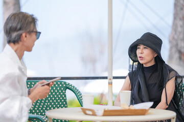 A Korean man and woman in their late 50s enjoy coffee and cake at a seaside cafe terrace with...
