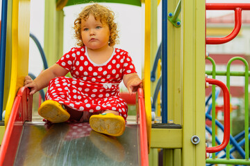 A young child wearing a red polka dot dress and yellow shoes plays happily on a playground slide The atmosphere is cheerful and colorful showcasing an enjoyable day outdoors