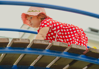 A young child wearing a red polka dot dress and a hat is climbing on colorful playground equipment The sun is shining in a vibrant outdoor park setting creating a joyful atmosphere