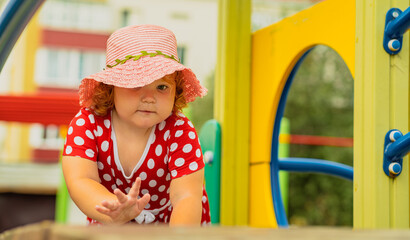 A young child wearing a red polka dot dress and a hat is climbing on colorful playground equipment The sun is shining in a vibrant outdoor park setting creating a joyful atmosphere