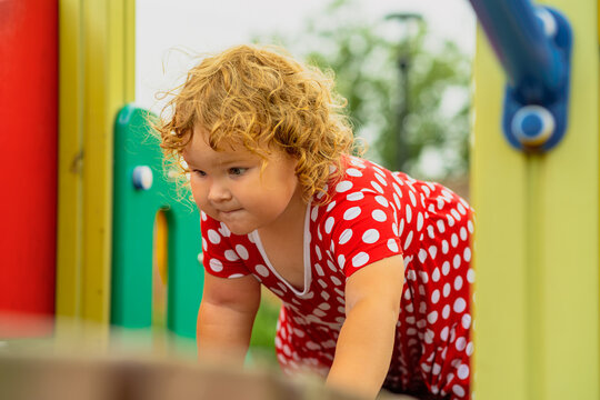 A young child wearing a red polka dot dress and a hat is climbing on colorful playground equipment The sun is shining in a vibrant outdoor park setting creating a joyful atmosphere