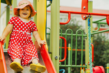 A happy kid in a red polka dot dress is sliding down a playground slide Bright colors surround her and the atmosphere is joyful during a sunny day with playful clouds