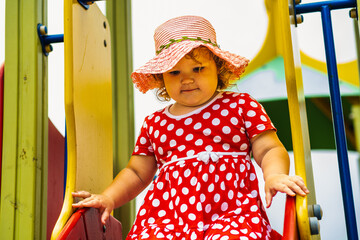 A happy kid in a red polka dot dress is sliding down a playground slide Bright colors surround her and the atmosphere is joyful during a sunny day with playful clouds