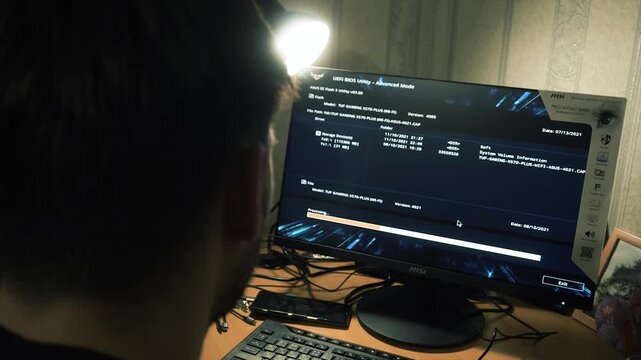 close-up. Blurred guy makes calculations looking at numbers, reports, program loading on computer screen while sitting with desk lamp. View from the headlines, back of the head. Apartment, night.