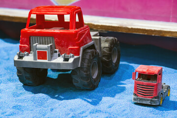 Two toy trucks are placed in a sandy play area A large red truck stands prominently next to a smaller red truck creating a playful contrast in size and inviting imaginative play for children
