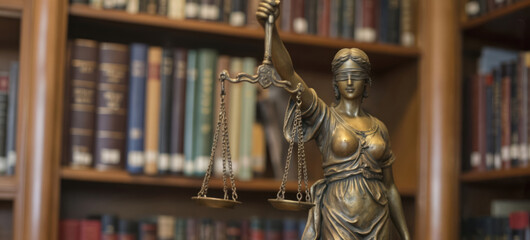Fototapeta premium Bronze sculpture of blindfolded Lady Justice holding scales stands in library. Colorful law books line shelves fairness, equality in legal system. Represents advocacy, justice, court, law, legal