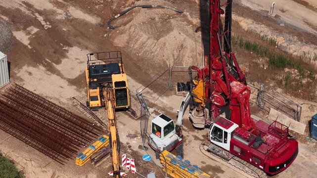Drone shot of a construction site featuring a red rotary drilling rig, excavator, steel reinforcement bars, and storage containers near a dirt road under development.