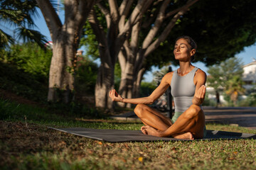 Serene middle-aged woman practicing seated meditation in lotus position outdoors at sunrise, eyes...
