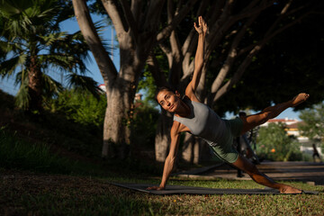 Strong athletic woman in her 40s performing a dynamic side plank pose with one arm and leg raised,...