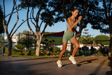 Fit athletic woman with brown hair jumping during a cardio workout in a sunny park, wearing green...
