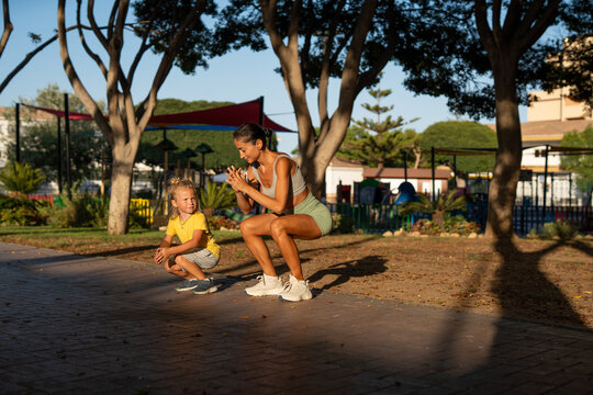 Fit young mother in sportswear doing squats outdoors while her little son watches and imitates her, both bonding and exercising together in a peaceful park during a sunny afternoon - Powered by Adobe