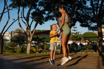 Joyful fit mother and her smiling little son jumping together during a workout session in the park,...