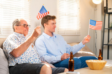 Happy family senior father and adult son holding USA flag enjoy watching and cheering sports competition live streaming on TV or celebration Independence Day together at home. Family relationship.