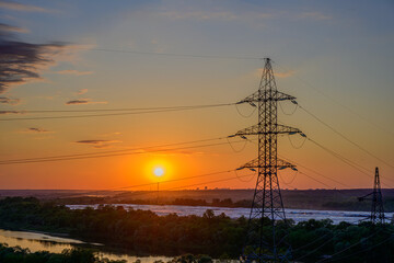 High-voltage power lines and electricity pylons at sunset over river and forest landscape, vivid orange sky, energy transmission infrastructure, industrial silhouettes, peaceful evening view