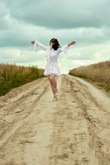 A young woman in a white dress with ruffles, stands with her arms outstretched on a dirt road,...