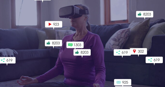 Sitting mature woman wearing VR headset meditating in living room, with floating social media icons - Powered by Adobe