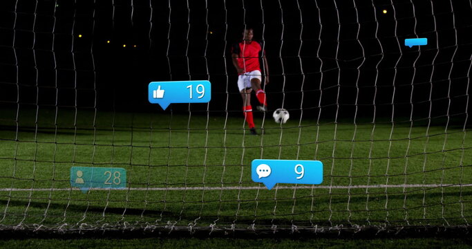 Kicking soccer player wearing red uniform on grass pitch at night, with net, ball, digital icons
