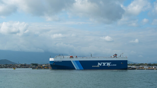 Large car carrier ship docked at port with overcast skies, mountains in the background, and vehicles lined up for transport