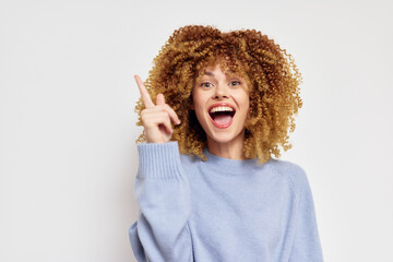Happy young woman with curly hair pointing upwards on a white background, wearing a light blue sweater, expressing excitement and joy Bright and cheerful concept ideal for positive vibes