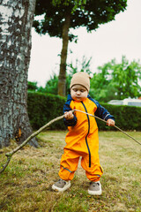 An Adventurous Toddler Dressed in a Bright Orange Rain Suit Joyfully Playing with a Long Stick in a...