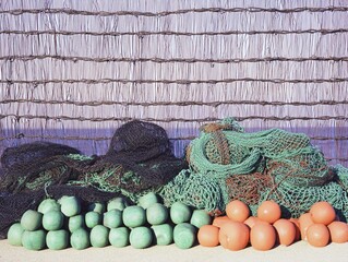 Fishing nets and buoys stacked against a woven reed fence in bright sunlight